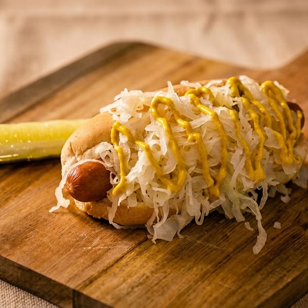 Hot dog with sauerkraut and mustard, served with a pickle spear on a wooden board.