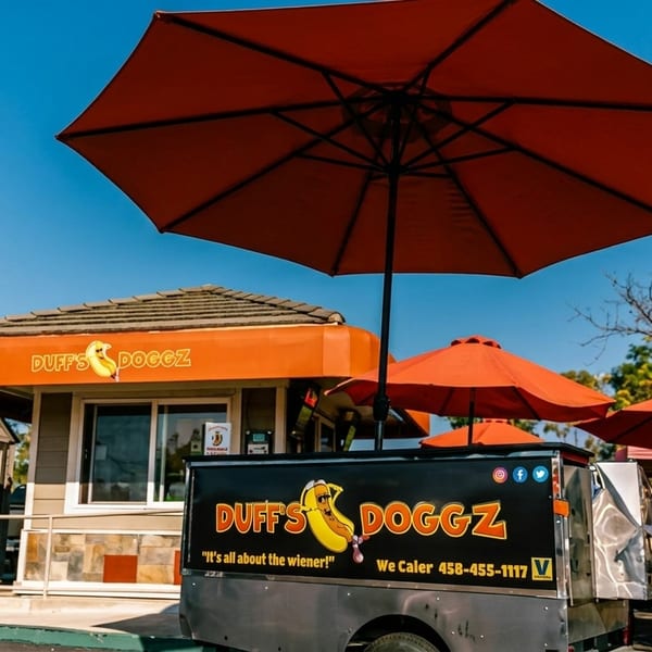 Duff's Doggz hot dog stand and food truck with orange umbrellas under a blue sky.