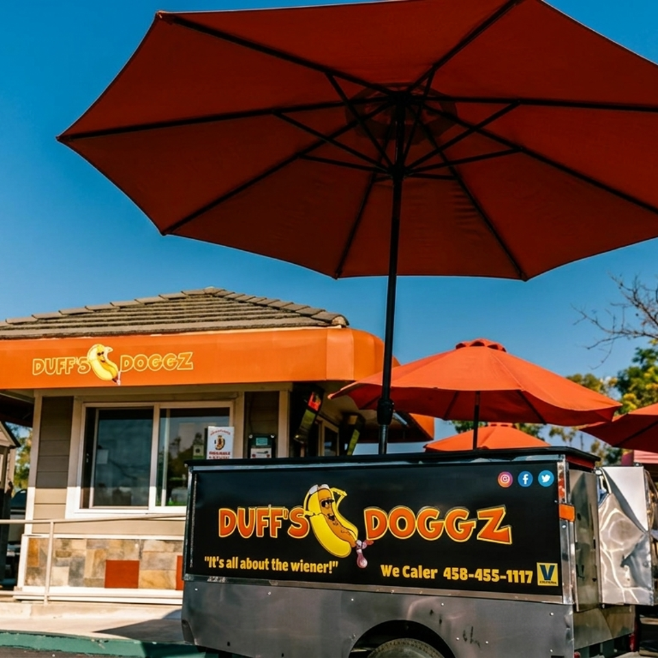 Duff's Doggz hot dog stand and food truck with orange umbrellas under a blue sky.