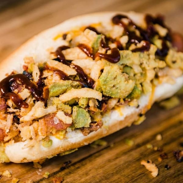 Gourmet hot dog topped with avocado, crispy onions, and BBQ sauce on a wooden surface.