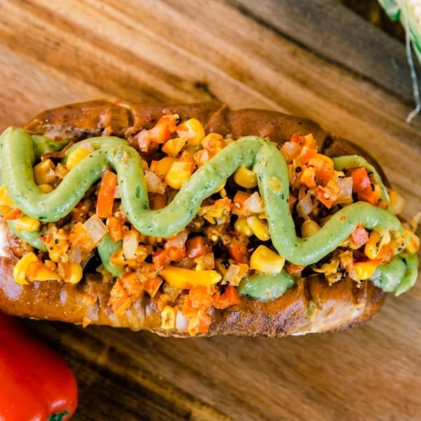 Gourmet hot dog topped with corn salsa and green sauce, alongside peppers and an ear of corn.